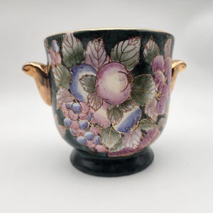Peut inclure: Un petit pot en céramique vert avec des poignées dorées. Le pot est décoré d'un motif floral avec des fleurs et des feuilles roses, violettes et bleues.
