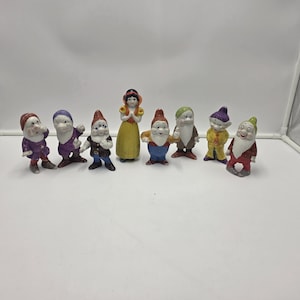 May include: A set of eight ceramic figurines featuring characters from "Snow White and the Seven Dwarfs." The collection includes Snow White in a yellow dress and seven dwarfs in colourful outfits. The figures are painted in bright colours.