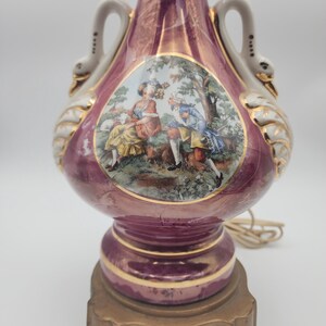 May include: A pink ceramic table lamp with swan-shaped handles and a gold base. The lamp features a painted scene of a couple in a garden.
