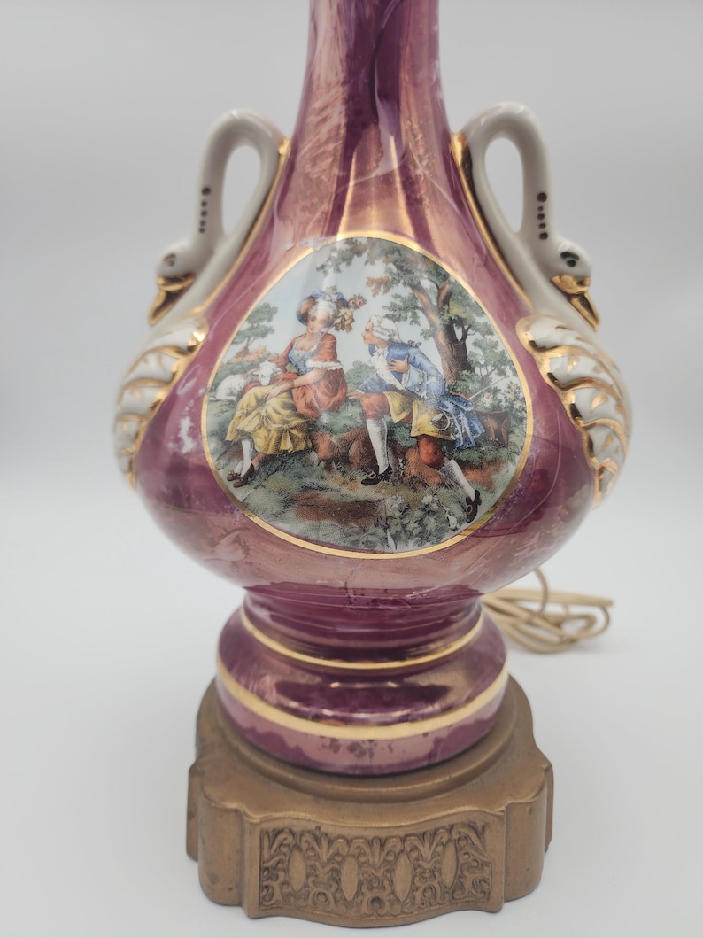 May include: A pink ceramic table lamp with swan-shaped handles and a gold base. The lamp features a painted scene of a couple in a garden.