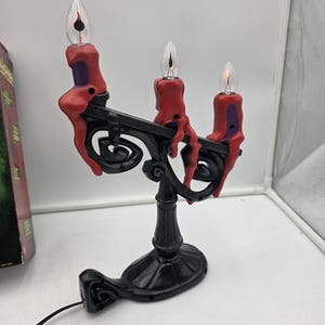 May include: A black candelabra lamp featuring three clear flame-shaped light bulbs topped with red wax-like drips. The base and arms are black with a swirling design, and the structure includes a black power cord extending from the bottom.