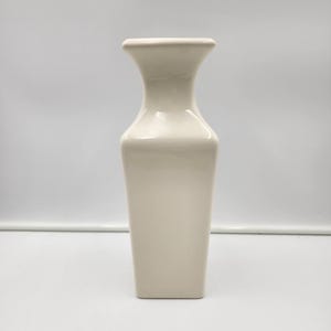 Könnte beinhalten: Eine hohe, cremefarbene Keramikvase mit quadratischem Boden und ausgestellter Oberseite. Die Vase hat eine glatte, glänzende Oberfläche und ein schlichtes, elegantes Design. Sie eignet sich zum Präsentieren von Blumen oder als dekoratives Element. Ca. 25 cm hoch.