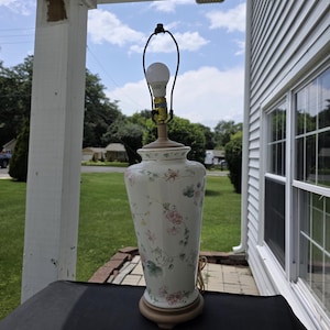 May include: A ceramic table lamp with a tapered, off-white body featuring a delicate floral pattern in shades of pink, green, and yellow. The lamp has a gold-toned metal harp, a light bulb, and a circular base in a muted, neutral brown tone.