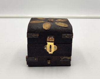 May include: A small, dark brown wooden box with a curved lid featuring gold-toned metal petal-shaped accents. The front includes a central gold-toned metal latch mechanism with a keyhole and horizontal metal strips along the sides of the box.