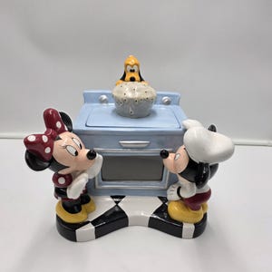 May include: A ceramic cookie jar shaped like a light blue kitchen stove with a black and white checkered base. Minnie Mouse stands on the left and a chef-hat-wearing mouse stands on the right. A small yellow dog figure sits on top of the stove.
