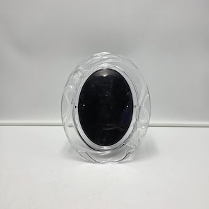 May include: An oval-shaped picture frame made of clear glass. The frame features a decorative, raised relief pattern of swirling lines and floral motifs around the perimeter. The center of the frame contains a solid black oval insert.