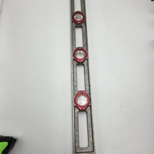 May include: A silver metal level with three red-rimmed bubble vials. The level is rectangular with open slots and is designed for measuring horizontal and vertical alignment. The vials are filled with a clear liquid and a bubble.