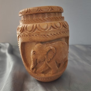 May include: A wooden carved vase with two elephants on the front. The vase is brown and has intricate carvings around the top.