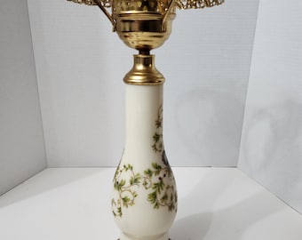 May include: A white ceramic table lamp with a gold metal base and shade. The lamp has a floral design with green leaves and blue flowers. The shade is a simple, round, openwork design.