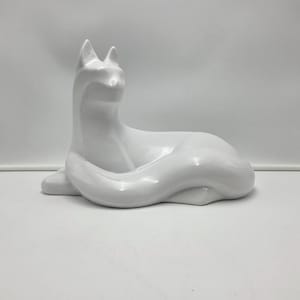 May include: A glossy white ceramic fox figurine in a relaxed, seated pose. The sculpture has a smooth, reflective surface and features detailed ears and a curled tail. The fox is a decorative item.