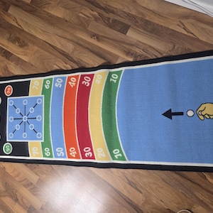May include: A rectangular game mat featuring a blue surface with a graphic of a hand and arrow, followed by multi-colored scoring zones labeled 10, 20, 30, 40, 50, 60, and 70. The black end section includes the word OUT and a grid target.