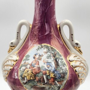 May include: A pink and white ceramic table lamp with gold accents. The lamp features a swan design with a scene of two people in a garden. The lamp has a gold base with an ornate design.