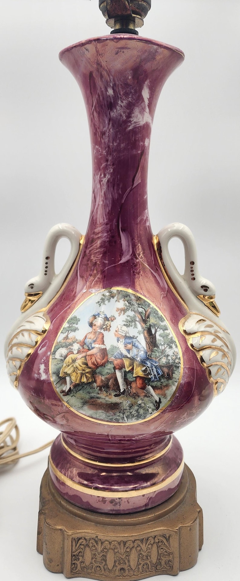 May include: A pink and white ceramic table lamp with gold accents. The lamp features a swan design with a scene of two people in a garden. The lamp has a gold base with an ornate design.