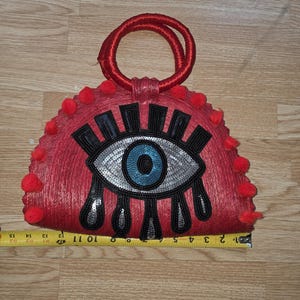 Puede incluir: Bolso de mano rojo con diseño de ojo. El bolso tiene un asa circular y está decorado con pompones rojos. El ojo está hecho de lentejuelas y adornos negros y plateados. El bolso mide aproximadamente 28 cm de ancho.