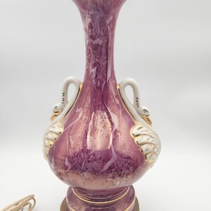 May include: A pink and white ceramic table lamp with gold accents. The lamp base features two swan handles and a marbled design. The lamp has a gold-colored cord.