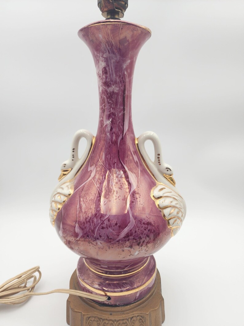 May include: A pink and white ceramic table lamp with gold accents. The lamp base features two swan handles and a marbled design. The lamp has a gold-colored cord.