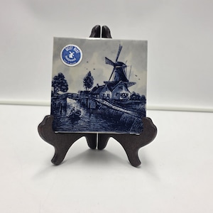 Op de afbeelding: Een Delft Blue tegel met een windmolen, gepresenteerd op een donkerbruin ezel. De tegel toont een traditioneel Nederlands landschap met een windmolen, een gracht en een boot. Een ronde sticker met tekst is zichtbaar.