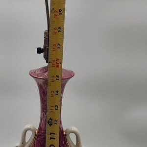 May include: A pink and white ceramic table lamp with gold accents. The lamp base features a swan design and a painted scene of people in a garden. The lamp is 20 inches tall.