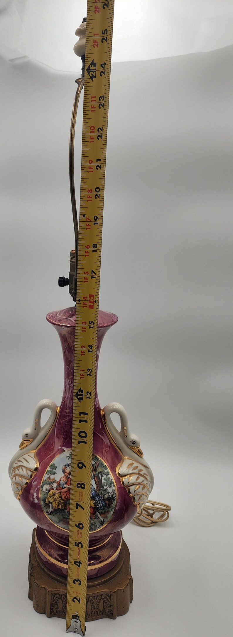 May include: A pink and white ceramic table lamp with gold accents. The lamp base features a swan design and a painted scene of people in a garden. The lamp is 20 inches tall.