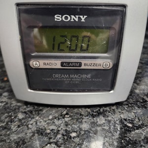 Peut inclure: Un radio-réveil Sony Dream Machine argenté avec un écran numérique affichant 12h00. Le radio-réveil possède des boutons pour la radio, l'alarme et la sonnerie.