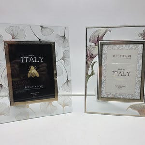 May include: Two decorative glass picture frames. One frame features a ginkgo leaf design, the other has calla lilies. Both frames have a gold-colored inner frame and the text "Made in ITALY" on the front.