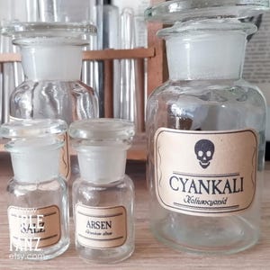 Może przedstawiać: Kolekcja zabytkowych szklanych butelek aptecznych z przezroczystymi szklanymi korkami. Butelki mają etykiety z tekstem i symbolem czaszki. Etykiety brzmią "CYANKALI", "ARSEN" i "THE SALE".