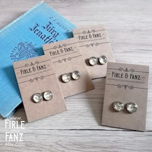 Op de afbeelding: Vier paar ronde oorknopjes met glazen cabochons met gedrukte tekst, elk bevestigd op individuele lichtbruine kartonnen displaykaarten met het label Firle & Fanz, gerangschikt bij een blauw boek met de titel Jürg Jenatsch.