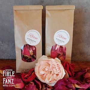 Op de afbeelding: Twee bruine papieren zakken met een doorzichtig venster waarin gedroogde rozenblaadjes te zien zijn. De zakken hebben witte etiketten met de tekst "Rosen" en "Blütenblätter" in zwart. De zakken staan op een houten oppervlak met een roze roos en rode rozenblaadjes.
