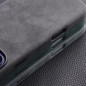 May include: Close-up of a dark gray phone case with a textured, suede-like finish. The case has a sleek design with cutouts for buttons and a camera. The case is likely designed to protect a smartphone.