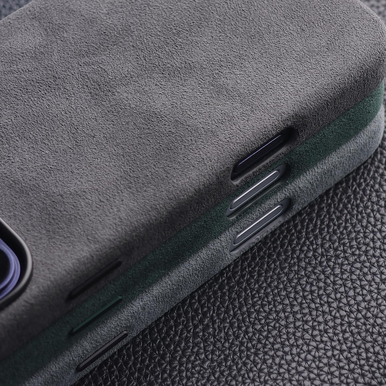 May include: Close-up of a dark gray phone case with a textured, suede-like finish. The case has a sleek design with cutouts for buttons and a camera. The case is likely designed to protect a smartphone.