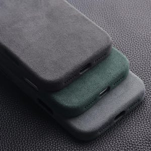 May include: Three phone cases in shades of gray and green are stacked on top of each other. The cases have a textured, suede-like finish. The cases are arranged diagonally, showcasing their side profiles and port openings.