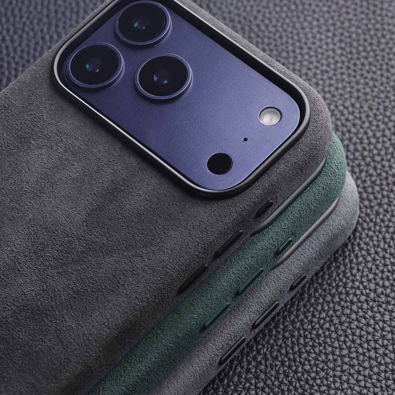 May include: Close-up of three phone cases in shades of gray and green. The cases have a textured, suede-like finish and are stacked, revealing the camera array of a phone. The top case is dark gray, the middle is green, and the bottom is light gray.