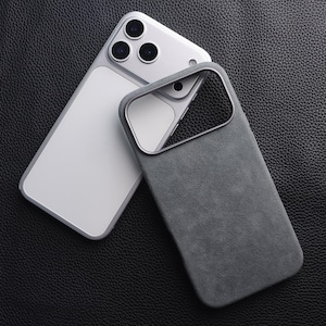 May include: A gray phone case with a soft, suede-like texture, next to a silver smartphone. The case has a camera cutout and a metallic border. The phone has three camera lenses.