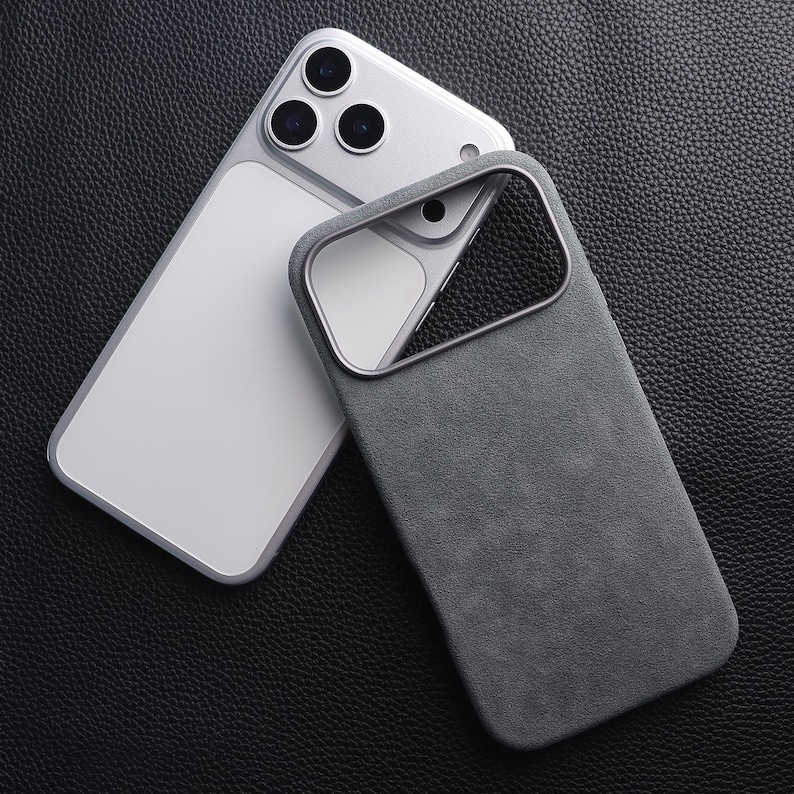 May include: A gray phone case with a soft, suede-like texture, next to a silver smartphone. The case has a camera cutout and a metallic border. The phone has three camera lenses.