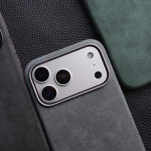 May include: Close-up of several phone cases in shades of gray and green. The cases have a matte finish and feature camera cutouts. The focus is on the details of the cases, showcasing their texture and design. The cases are arranged on a textured surface.