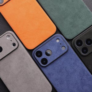 K&ouml;nnte beinhalten: Draufsicht auf mehrere bunte Handyh&uuml;llen. Die H&uuml;llen sind in Orange-, Gr&uuml;n-, Blau-, Grau- und Schwarzt&ouml;nen gehalten. Jede H&uuml;lle hat eine strukturierte, Wildleder-&auml;hnliche Oberfl&auml;che und Kameraausschnitte.