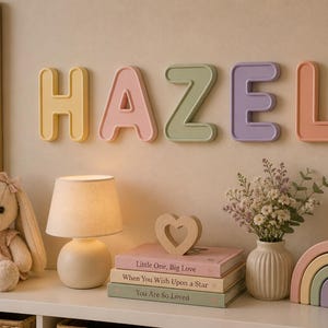 May include: A set of five individual wall-mounted letters spelling the name HAZEL in pastel colours including yellow, pink, sage green, lavender, and peach. Each letter features a rounded, raised border design, creating a soft and dimensional appearance.