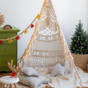 Op de afbeelding: Een witte macramé tipi met lichtsnoeren erin, twee grijze kussens en een wit tapijt. De tipi is versierd met een guirlande van houten kralen en kleurrijke kwastjes.