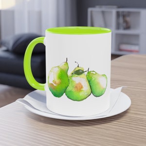 Könnte beinhalten: Eine weiße Keramiktasse mit grünem Rand und Henkel. Die Tasse zeigt eine Aquarell-Illustration von drei grünen Birnen.