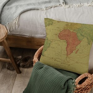 Può includere: Un cuscino decorativo con un design di mappa del mondo vintage. La mappa è stampata su un tessuto beige e presenta una combinazione di colori rosso e arancione. Il cuscino è seduto in un cesto di vimini.