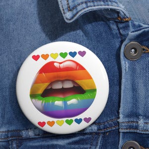 May include: A white button with a rainbow-colored open mouth design. The lips are painted in the colors of the rainbow, with a row of colorful hearts above and below the mouth. The button is pinned to a denim jacket.