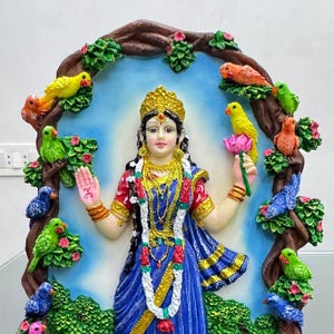 Puede incluir: Una estatua colorida de una diosa hindú con un sari azul, rodeada de pájaros en un árbol. La estatua está pintada en colores vibrantes y tiene un diseño detallado.