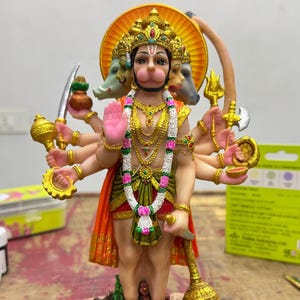 Puede incluir: Estatua detallada de una deidad con múltiples brazos y rostro de mono, adornada con joyas de oro y un halo. La figura viste ropas rojas y verdes y se alza sobre una base decorativa. Sostiene varios objetos simbólicos.