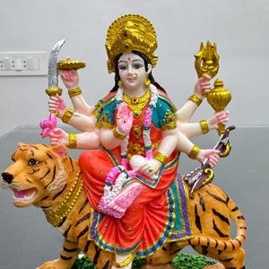 Puede incluir: Una estatua colorida de una diosa hindú montando un tigre. La diosa tiene múltiples brazos y está adornada con joyas. El tigre es marrón y naranja con rayas negras. La estatua está colocada sobre una base blanca con un borde negro.
