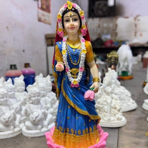 Könnte beinhalten: Eine Figur einer Frau in einem blauen und goldenen Sari, die auf einer rosa Lotusblume steht. Sie trägt eine Blumenkette und hält eine rosa Lotusblume in der Hand.