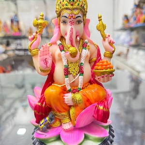 May include: A colourful figurine of the Hindu deity Ganesha, seated on a pink lotus flower. He is depicted with an elephant head, four arms, and a large belly. He is wearing a gold crown, a red and orange robe, and a garland of flowers. He is holding a sweet treat in one hand and a small axe in another. A small rat is sitting at his feet.