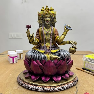 May include: A detailed statue of a deity with multiple faces and arms, seated on a lotus flower base. The statue is painted with gold, purple, and bronze tones. The deity holds various objects, including a flower and a small container.