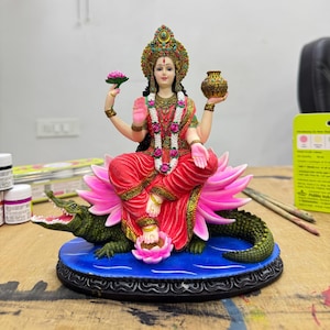 May include: A detailed statue of a seated deity, adorned in red and gold garments, holding a lotus flower and a pot. The figure is seated on a pink lotus flower, atop a crocodile, with a blue base. The statue has intricate details and vibrant colors.