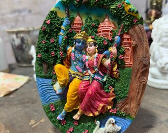 May include: A colorful 3D wall hanging depicting a Hindu deity couple, Radha and Krishna, on a swing in a lush green forest setting. The scene includes a river, flowers, birds, and a temple in the background.