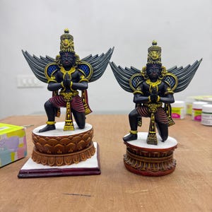 May include: Two detailed statues of a deity with black skin, gold accents, and large, dark wings. The figures are kneeling with hands clasped in prayer, wearing ornate headdresses and patterned clothing. They stand on decorative, tiered bases.